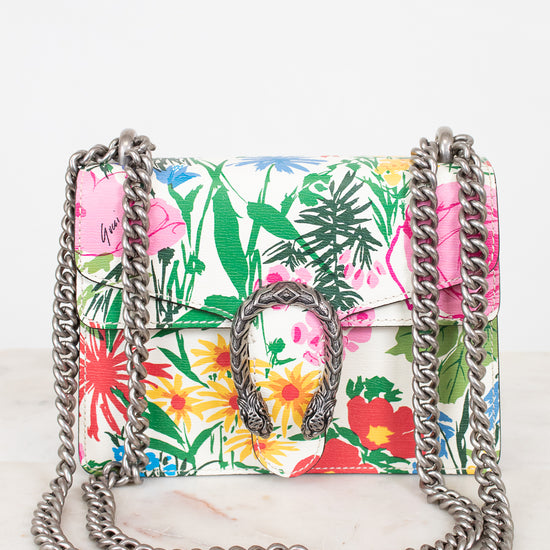 A small structured handbag with a vibrant tropical floral print in pink, green, yellow, and red on a white base, featuring a silver-tone metal chain strap and a central metal dionysus closure.