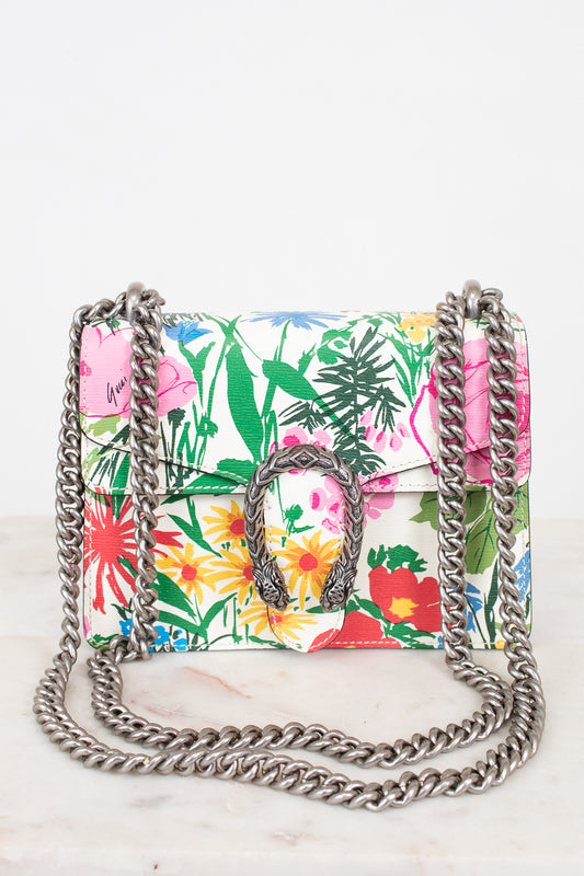 A small structured handbag with a vibrant tropical floral print in pink, green, yellow, and red on a white base, featuring a silver-tone metal chain strap and a central metal dionysus closure.