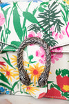 A close-up of a Gucci flap bag featuring a vibrant tropical floral print with green palm leaves, pink flowers, and yellow sunflowers on a white base, accented by a prominent silver-tone dionysus closure.