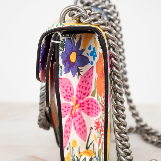 A close-up of a handbag with a colorful floral pattern and a chunky silver chain strap, set against a clean white background.