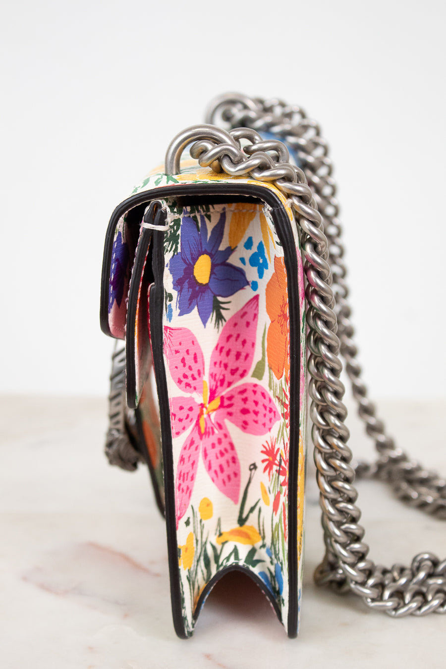 A close-up of a handbag with a colorful floral pattern and a chunky silver chain strap, set against a clean white background.