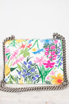 A rectangular shoulder bag with a vibrant multicolor floral print on a white background, featuring a silver-tone metal chain strap, photographed on a light marble surface against a white background.