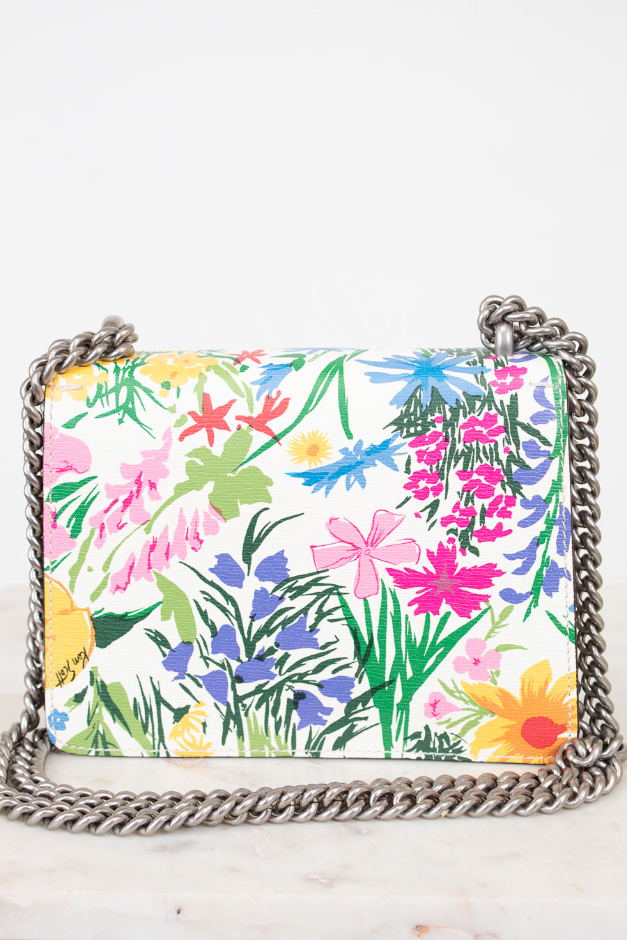 A rectangular shoulder bag with a vibrant multicolor floral print on a white background, featuring a silver-tone metal chain strap, photographed on a light marble surface against a white background.