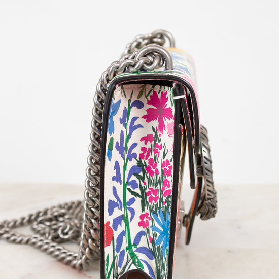 A close-up side view of a handbag with a colorful floral pattern on the front panel and a silver chain strap, resting on a light marble surface against a white background.