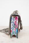 A close-up side view of a handbag with a colorful floral pattern on the front panel and a silver chain strap, resting on a light marble surface against a white background.