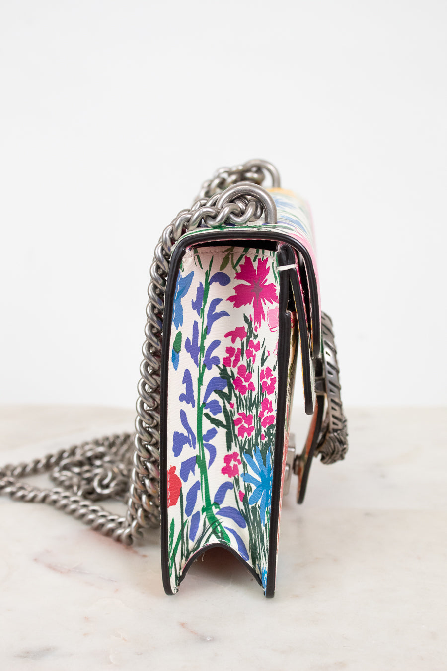 A close-up side view of a handbag with a colorful floral pattern on the front panel and a silver chain strap, resting on a light marble surface against a white background.