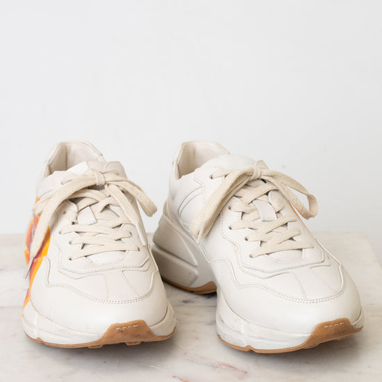 A pair of chunky low-top sneakers in off-white leather with beige laces and an orange and yellow graphic accent on the side, resting on a light marble surface against a white background.