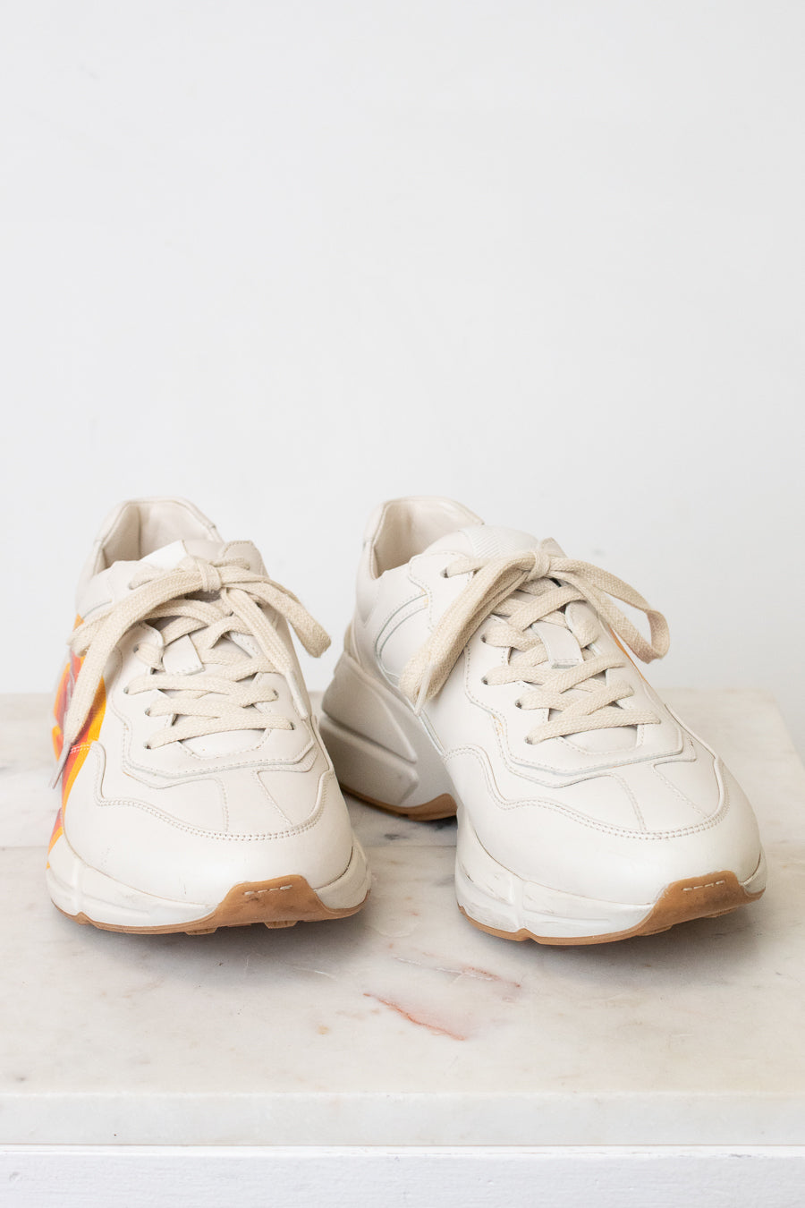 A pair of chunky low-top sneakers in off-white leather with beige laces and an orange and yellow graphic accent on the side, resting on a light marble surface against a white background.