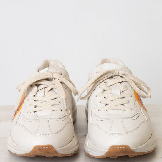 A white leather sneaker featuring a red, orange, and yellow graphic on the side with a stylized interlocking G logo, set against a plain white background.