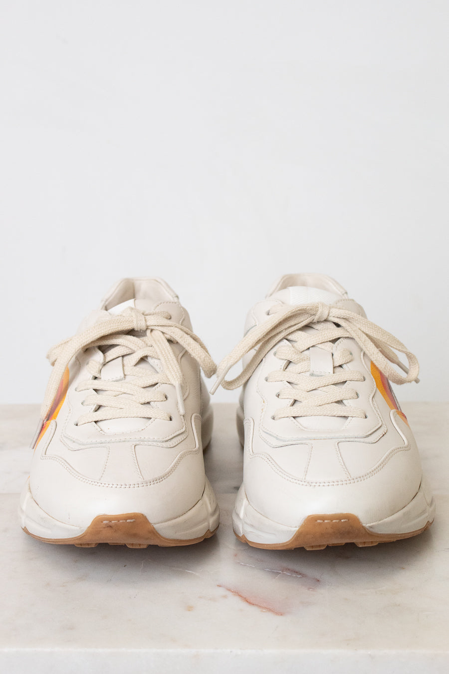 A white leather sneaker featuring a red, orange, and yellow graphic on the side with a stylized interlocking G logo, set against a plain white background.