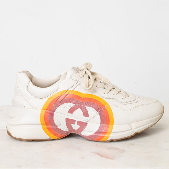 A white leather sneaker featuring a large circular red, orange, and yellow graphic on the side with a stylized interlocking G logo, set against a plain white background.