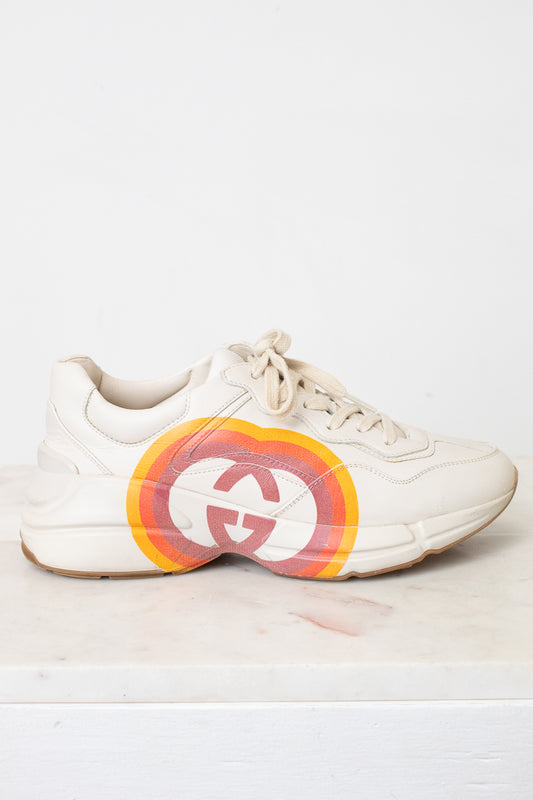 A white leather sneaker featuring a large circular red, orange, and yellow graphic on the side with a stylized interlocking G logo, set against a plain white background.