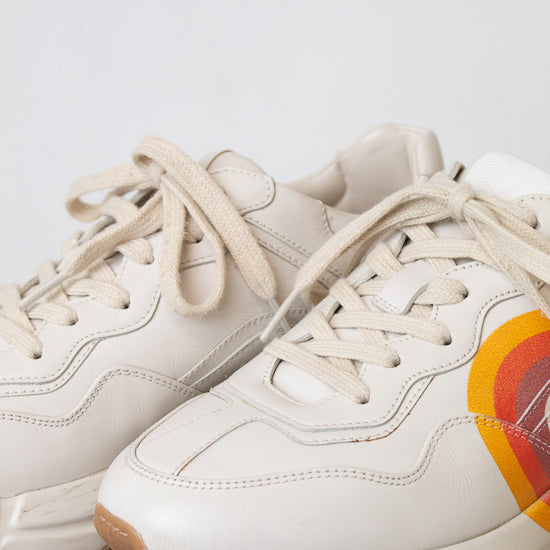 A white leather sneaker featuring a red, orange, and yellow graphic on the side with a stylized interlocking G logo, set against a plain white background.