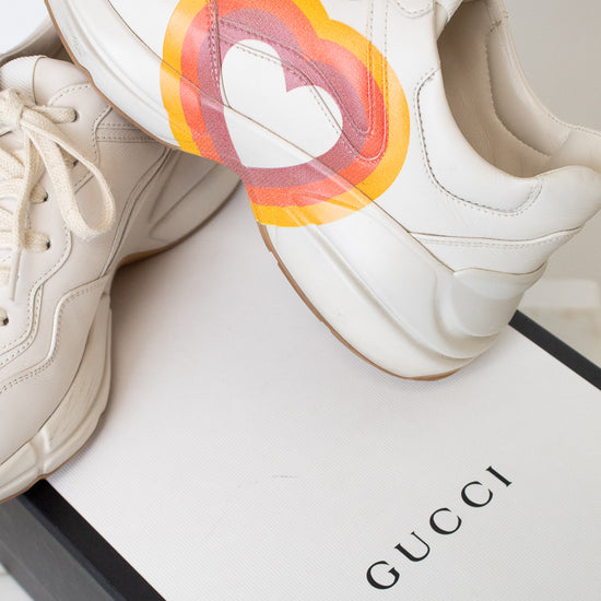 A white leather sneaker featuring a large circular red, orange, and yellow graphic on the side with a stylized interlocking G logo, set against a white and black "Gucci" box