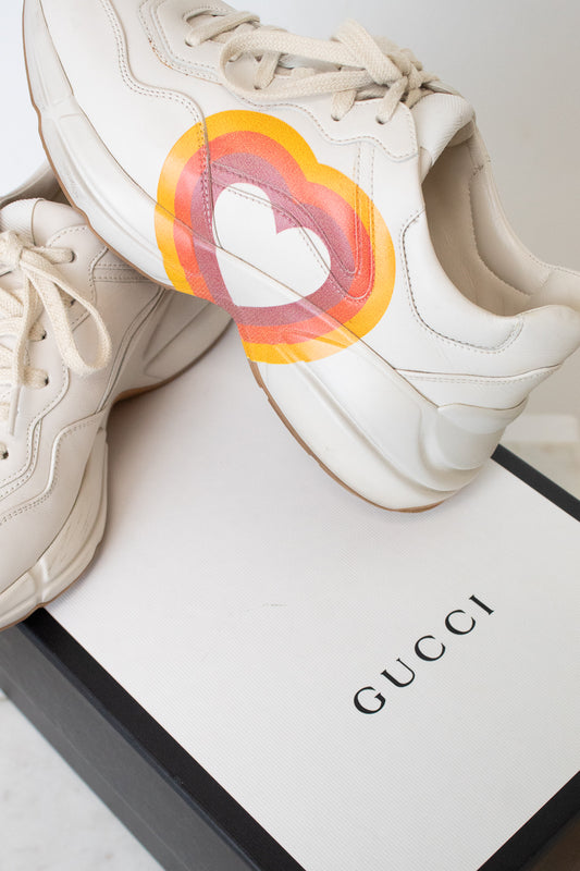 A white leather sneaker featuring a large circular red, orange, and yellow graphic on the side with a stylized interlocking G logo, set against a white and black "Gucci" box