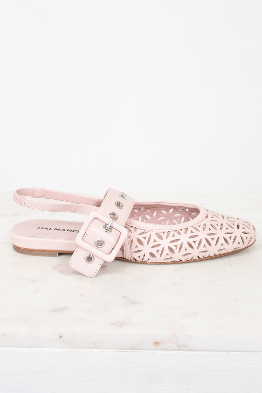 A single light pink flat shoe with a geometric laser-cut pattern on the toe and a matching strap with a buckle closure, shown on a white marble surface against a white background.