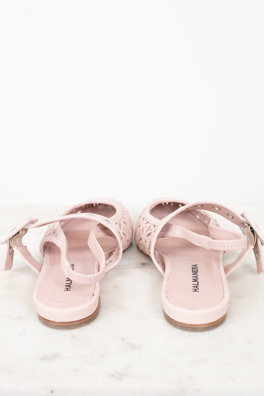 A pair of light pink flat sandals with cutout details and adjustable straps, shown from the back on a white marble surface against a white background.
