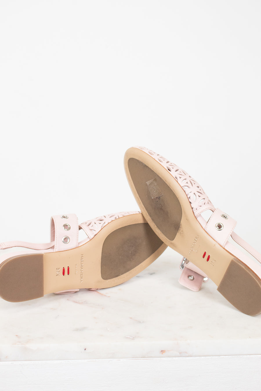 A pair of light pink leather flat sandals with laser-cut geometric patterns on the toe straps, displayed on a white marble surface against a white background.