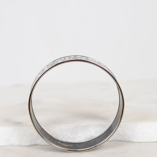 A silver-toned metal bangle bracelet with a textured, enamel and pattern on the outer surface, displayed on a white marble surface against a white background.