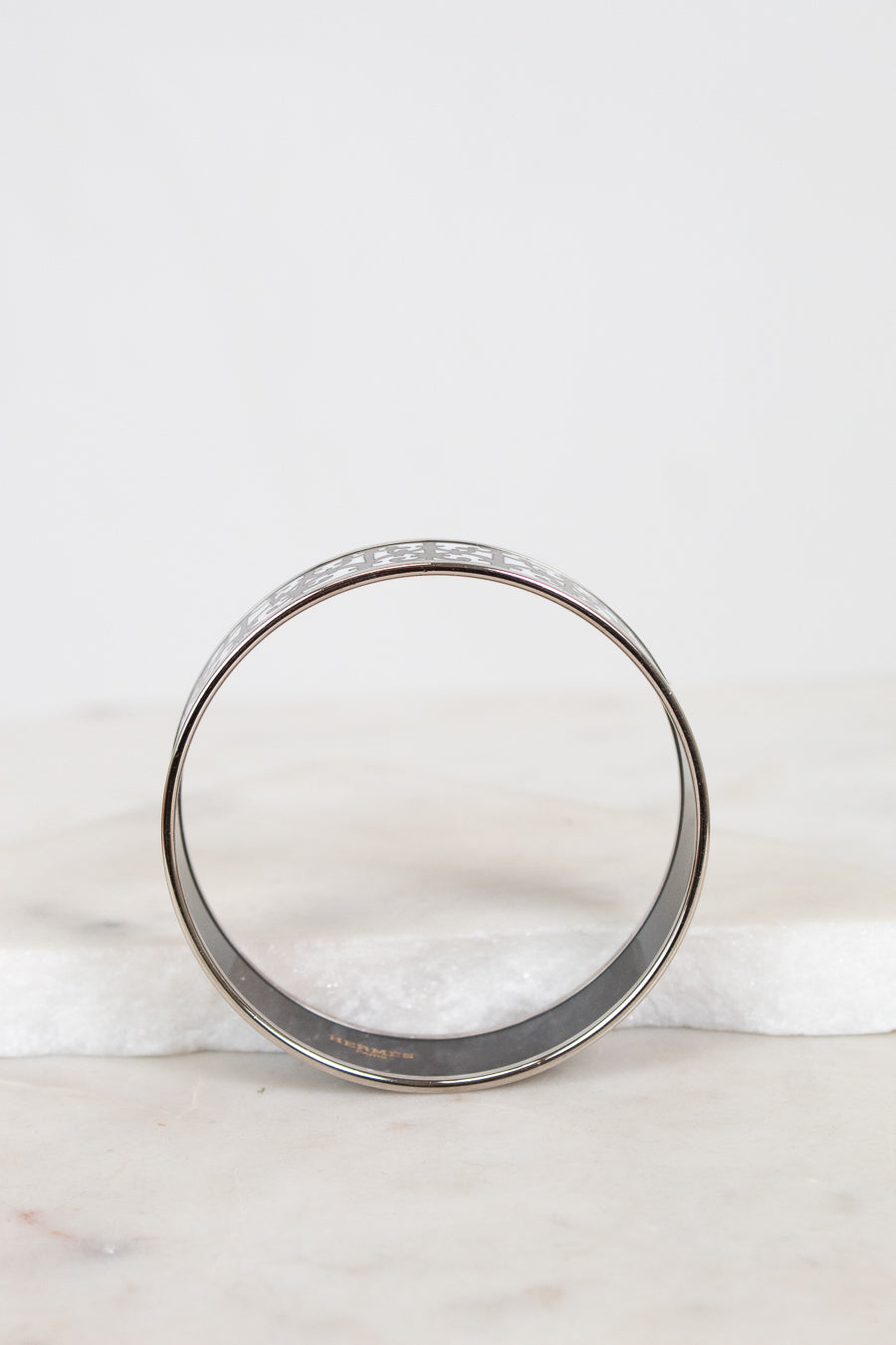 A silver-toned metal bangle bracelet with a textured, enamel and pattern on the outer surface, displayed on a white marble surface against a white background.