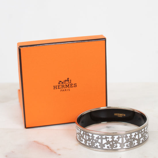 An Hermès logo bangle bracelet in silver-tone metal, displayed in front of its signature orange Hermès Paris box on a light marble surface.