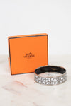 An Hermès logo bangle bracelet in silver-tone metal, displayed in front of its signature orange Hermès Paris box on a light marble surface.