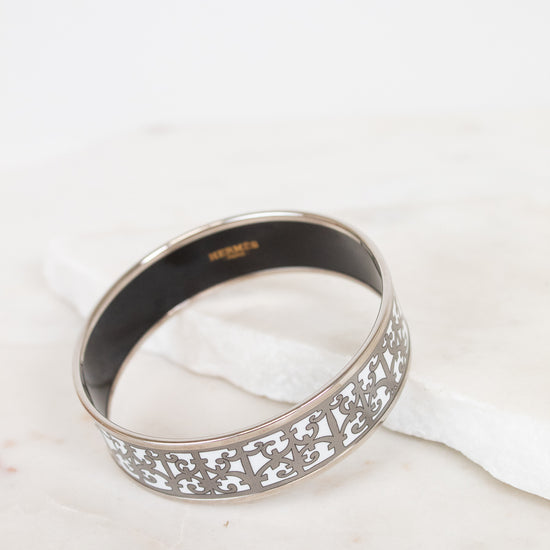 A silver-toned metal bangle bracelet with an intricate openwork filigree pattern on the outer band and a smooth black interior, resting on a white marble surface.