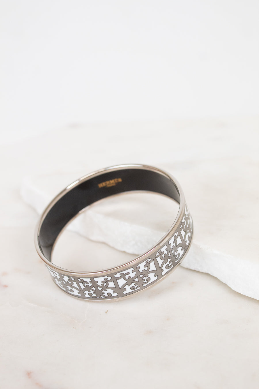 A silver-toned metal bangle bracelet with an intricate openwork filigree pattern on the outer band and a smooth black interior, resting on a white marble surface.