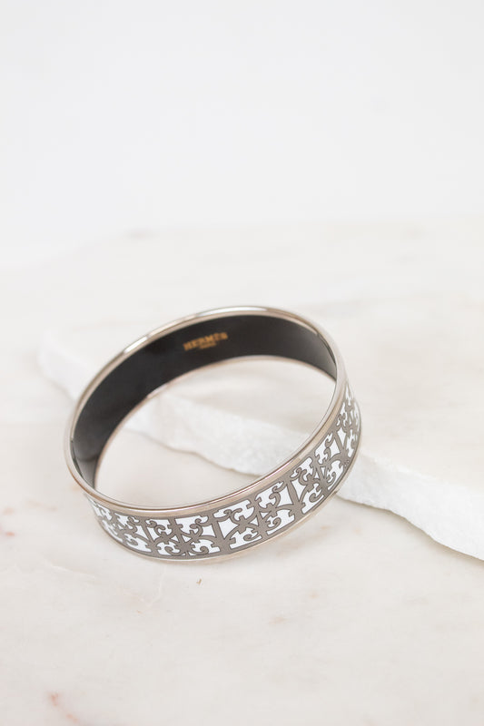 A silver-toned metal bangle bracelet with an intricate openwork filigree pattern on the outer band and a smooth black interior, resting on a white marble surface.