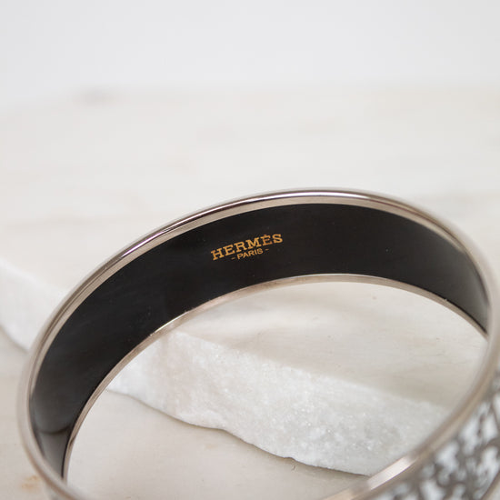 A close-up of a Hermès bangle bracelet with a black enamel interior and a silver-tone outer band featuring a white and gray patterned design.