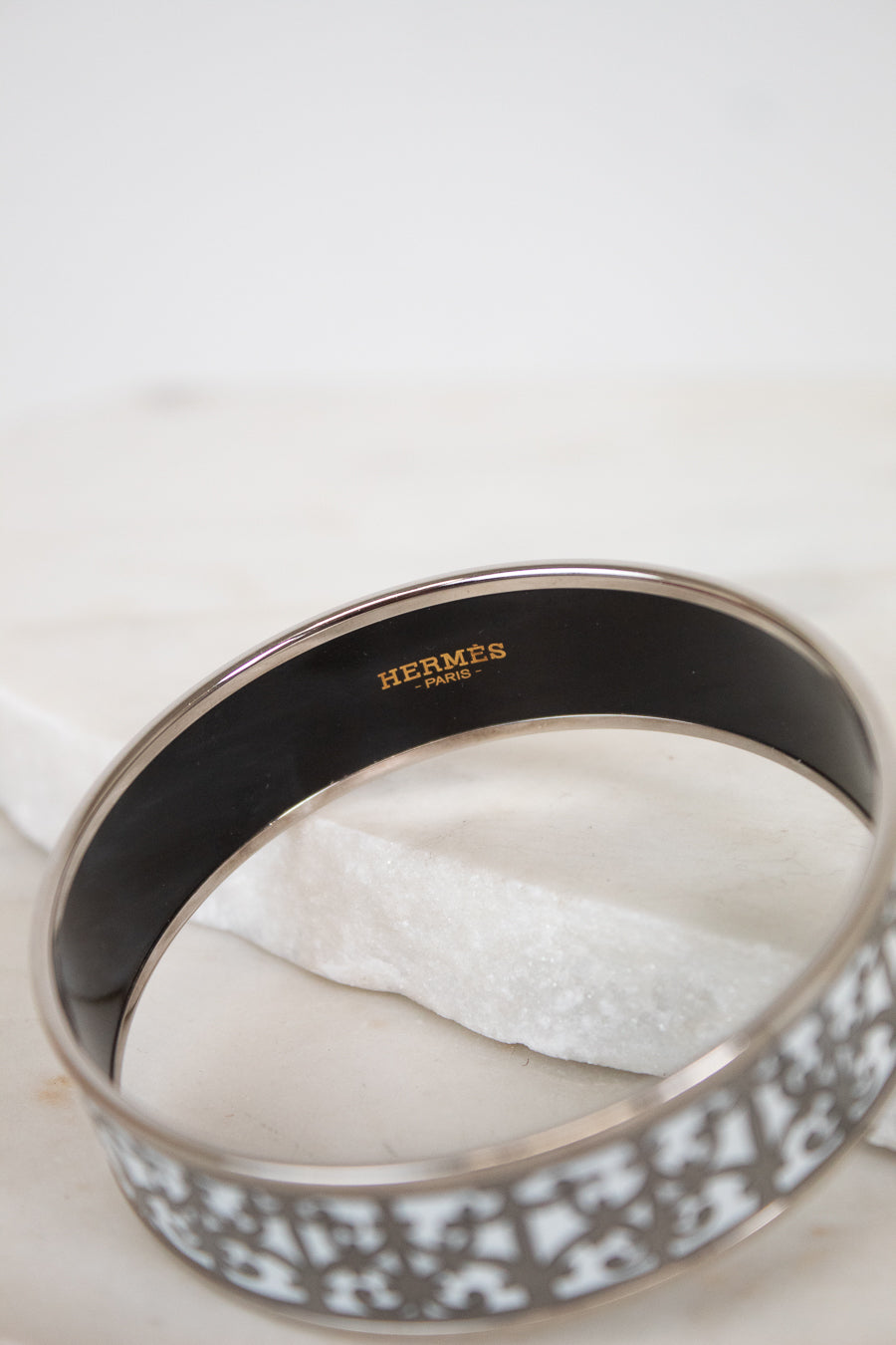 A close-up of a Hermès bangle bracelet with a black enamel interior and a silver-tone outer band featuring a white and gray patterned design.