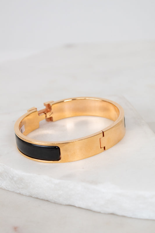 A gold-tone hinge bracelet with a smooth black inlay, displayed on a white marble slab against a soft, light gray background.