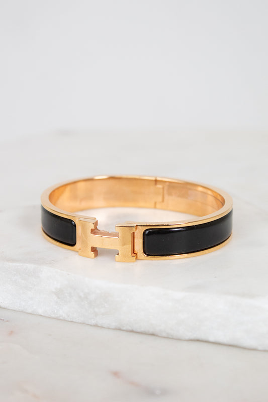 A gold-tone hinged cuff bracelet with a glossy black enamel inlay, resting on a white marble slab against a light gray background.