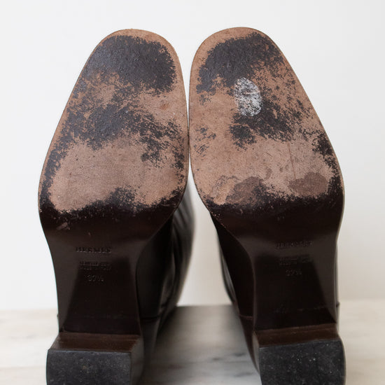 A pair of dark brown leather boots shown from the sole, revealing heavily worn and scuffed soles with visible scuffing and a white marking on the upper portions.