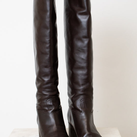 A pair of dark brown leather over-the-knee boots with block heels, displayed on a light-colored stone surface against a plain off-white background.