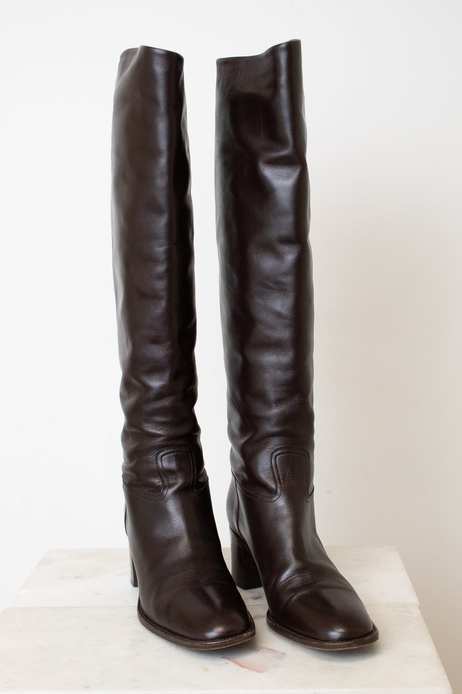A pair of dark brown leather over-the-knee boots with block heels, displayed on a light-colored stone surface against a plain off-white background.