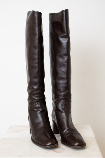 A pair of dark brown leather over-the-knee boots with block heels, displayed on a light-colored stone surface against a plain off-white background.