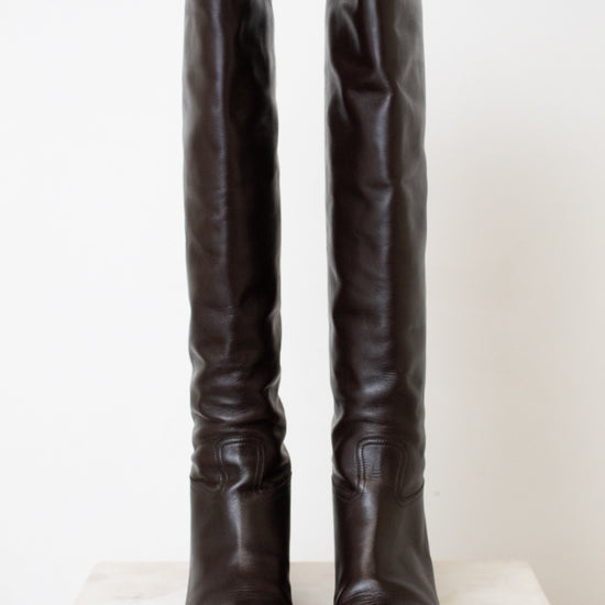 A pair of dark brown leather over-the-knee boots with a slightly slouchy fit and low block heels, displayed on a light-colored stone surface against a plain off-white background.