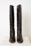 A pair of dark brown leather over-the-knee boots with a slightly slouchy fit and low block heels, displayed on a light-colored stone surface against a plain off-white background.
