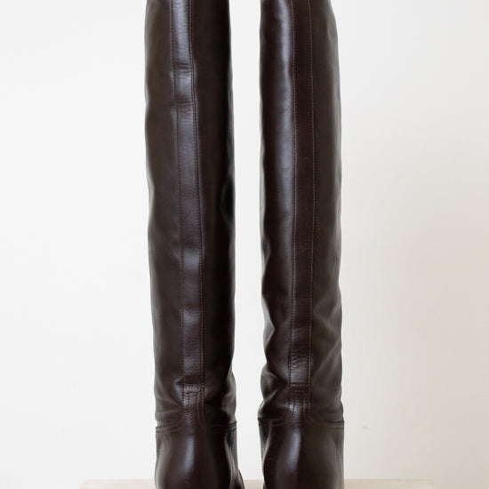 A pair of dark brown leather over-the-knee boots shown from the back, standing upright on a light-colored stone surface against a plain off-white background.