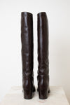 A pair of dark brown leather over-the-knee boots shown from the back, standing upright on a light-colored stone surface against a plain off-white background.
