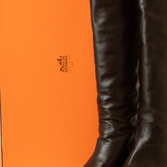 A pair of dark brown leather knee-high boots standing in front of a bright orange Hermès box, with the Hermès logo visible on the packaging.