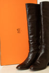 A pair of dark brown leather knee-high boots standing in front of a bright orange Hermès box, with the Hermès logo visible on the packaging.