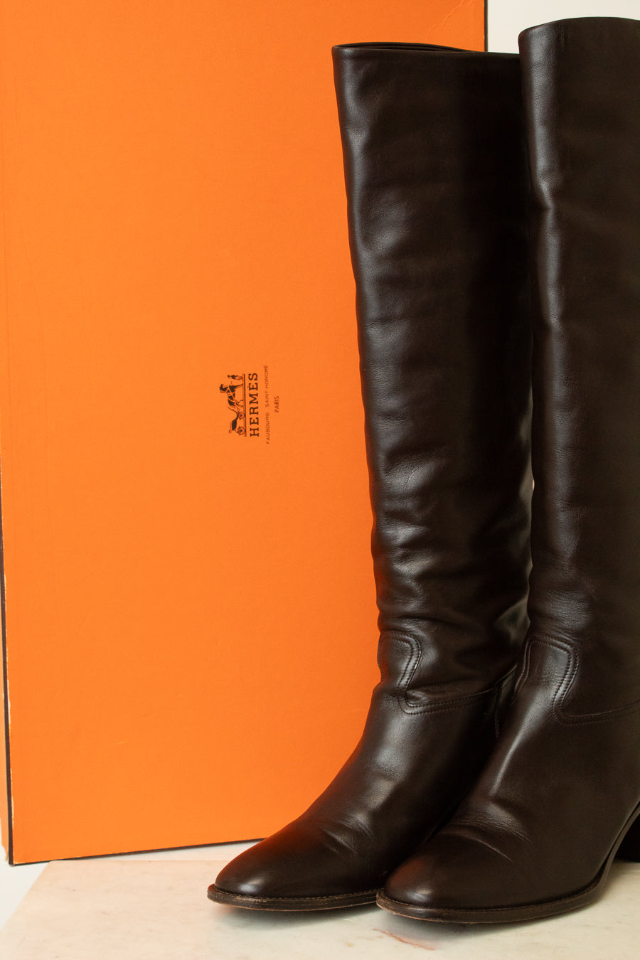 A pair of dark brown leather knee-high boots standing in front of a bright orange Hermès box, with the Hermès logo visible on the packaging.