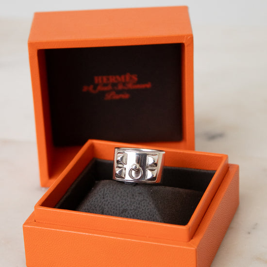 Silver HERMES ring with a geometric design and central circular detail, displayed in an orange HERMES jewelry box on a white background.