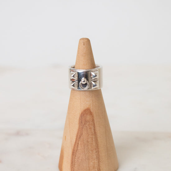 Silver HERMES ring with a wide band, geometric cutout details, and a central circular motif, displayed on a wooden ring stand.The ring is placed on a wooden ring stand with a white background.