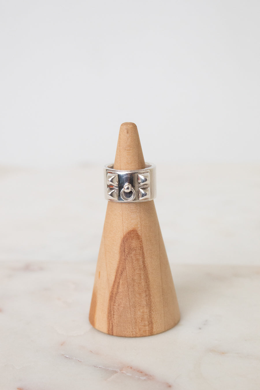 Silver HERMES ring with a wide band, geometric cutout details, and a central circular motif, displayed on a wooden ring stand.The ring is placed on a wooden ring stand with a white background.