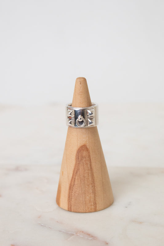 Silver HERMES ring with a wide band, geometric cutout details, and a central circular motif, displayed on a wooden ring stand.The ring is placed on a wooden ring stand with a white background.