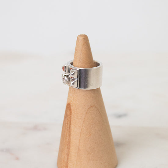 Side view of a silver HERMES ring featuring a wide polished band, geometric cutouts, and a raised circular emblem in the middle of the ring. The ring is resting on a wooden ring holder on a marble background.