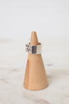Side view of a silver HERMES ring featuring a wide polished band, geometric cutouts, and a raised circular emblem in the middle of the ring. The ring is resting on a wooden ring holder on a marble background.